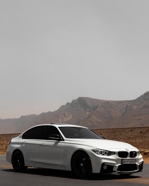 A sleek white sedan is parked on a deserted road with a mountainous landscape in the background. The car has tinted windows and a sporty design, emphasizing its modern appeal. The road stretches into the distance, suggesting a sense of freedom and adventure.
