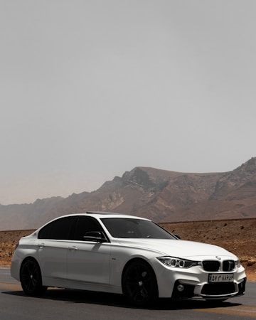 A sleek white sedan is parked on a deserted road with a mountainous landscape in the background. The car has tinted windows and a sporty design, emphasizing its modern appeal. The road stretches into the distance, suggesting a sense of freedom and adventure.
