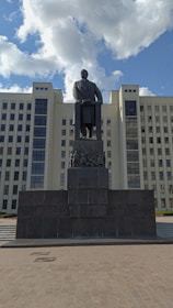 A tall statue of a man stands prominently in front of a large, multi-windowed government building with classical architecture. The sky is partly cloudy, providing a backdrop of fluffy white clouds against a blue sky.