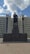 A tall statue of a man stands prominently in front of a large, multi-windowed government building with classical architecture. The sky is partly cloudy, providing a backdrop of fluffy white clouds against a blue sky.