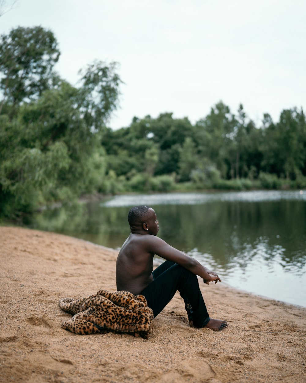 낮 동안 호수 근처의 갈색 모래 위에 앉아 있는 검은 반바지를 입은 소년