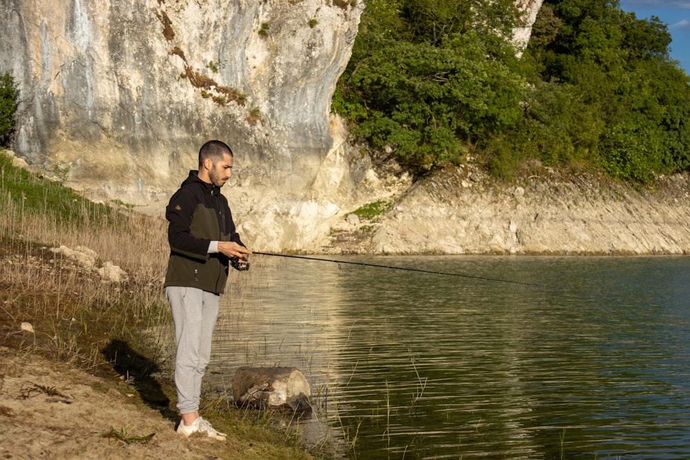 uomo in giacca nera e pantaloni bianchi in piedi sulla roccia vicino allo specchio d'acqua durante il giorno