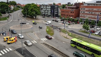 An urban intersection with multiple vehicles including cars, a yellow van, and a green bus. Pedestrians are visible walking near the intersection. The area is surrounded by buildings, including a brick commercial building. Some signs and banners are present near the road. Green trees and grass are seen in certain areas.