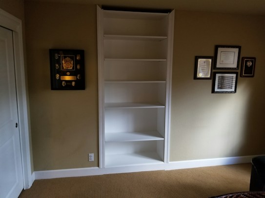 A room features an empty white built-in shelf set against a beige wall with several framed awards and certificates displayed around it. The floor is covered with light brown carpet, and there is partial view of white doors to the left and a dark piece of furniture on the right.
