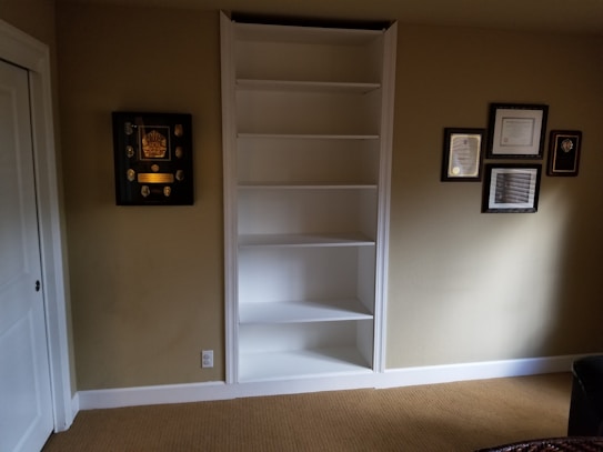 A room features an empty white built-in shelf set against a beige wall with several framed awards and certificates displayed around it. The floor is covered with light brown carpet, and there is partial view of white doors to the left and a dark piece of furniture on the right.
