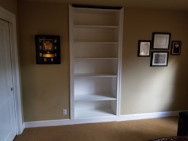 A room features an empty white built-in shelf set against a beige wall with several framed awards and certificates displayed around it. The floor is covered with light brown carpet, and there is partial view of white doors to the left and a dark piece of furniture on the right.