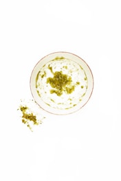 A bowl of yogurt with a green herb topping, possibly pesto or chimichurri, sits in the center. Small specks of the green mixture are visible around the bowl on a white surface.