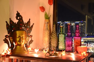 A decorative setup featuring an ornate golden Buddha head with intricate detailing, surrounded by colorful glass bottles in green, purple, and red. The scene is illuminated by warm string lights and adorned with two red tulips in a glass vase nearby, set against a background of a cityscape at night.