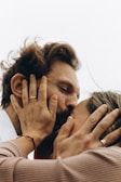 A close-up shot of two people sharing an intimate moment, with their hands gently holding each other's faces. Both individuals are wearing rings, and one has a red string bracelet on their wrist. The image conveys a sense of closeness and affection.