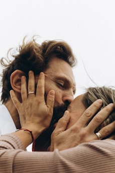 A close-up shot of two people sharing an intimate moment, with their hands gently holding each other's faces. Both individuals are wearing rings, and one has a red string bracelet on their wrist. The image conveys a sense of closeness and affection.