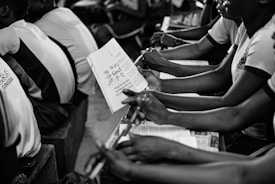 A group of students seated closely together in what appears to be a classroom setting, holding notebooks and pens. They are attentively engaged in what seems like a lecture or group discussion. The focus is on the students' hands and the open notebook, where notes are visible.