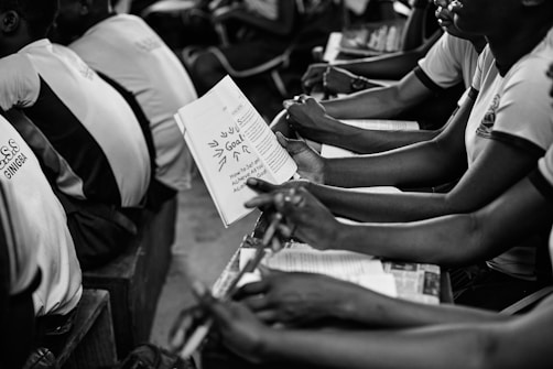 A group of students seated closely together in what appears to be a classroom setting, holding notebooks and pens. They are attentively engaged in what seems like a lecture or group discussion. The focus is on the students' hands and the open notebook, where notes are visible.