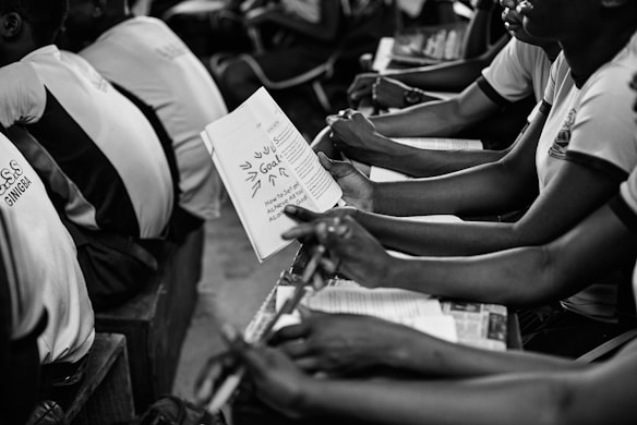A group of students seated closely together in what appears to be a classroom setting, holding notebooks and pens. They are attentively engaged in what seems like a lecture or group discussion. The focus is on the students' hands and the open notebook, where notes are visible.