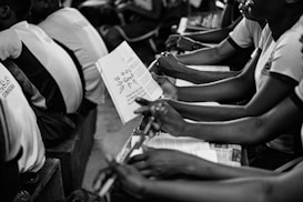 A group of students seated closely together in what appears to be a classroom setting, holding notebooks and pens. They are attentively engaged in what seems like a lecture or group discussion. The focus is on the students' hands and the open notebook, where notes are visible.