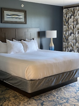 A neatly made bed with white linens and four pillows is placed against a dark wooden headboard. There is an abstract painting with a wooden frame hung above the headboard. A modern table lamp with a blue and white gradient base stands beside the bed. The room features a patterned carpet and curtains with a leaf motif.