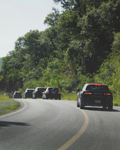 A convoy of covered car carriers driving along a scenic highway in India.