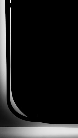 A minimalistic black and white image with a large black shape occupying most of the space. The shape has smooth, curved edges and is bordered by a narrow white strip. The contrast between the black and white creates a dramatic visual effect.