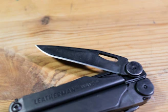 A sharp multitool unfolded on a wooden picnic table with sunlight filtering through trees.