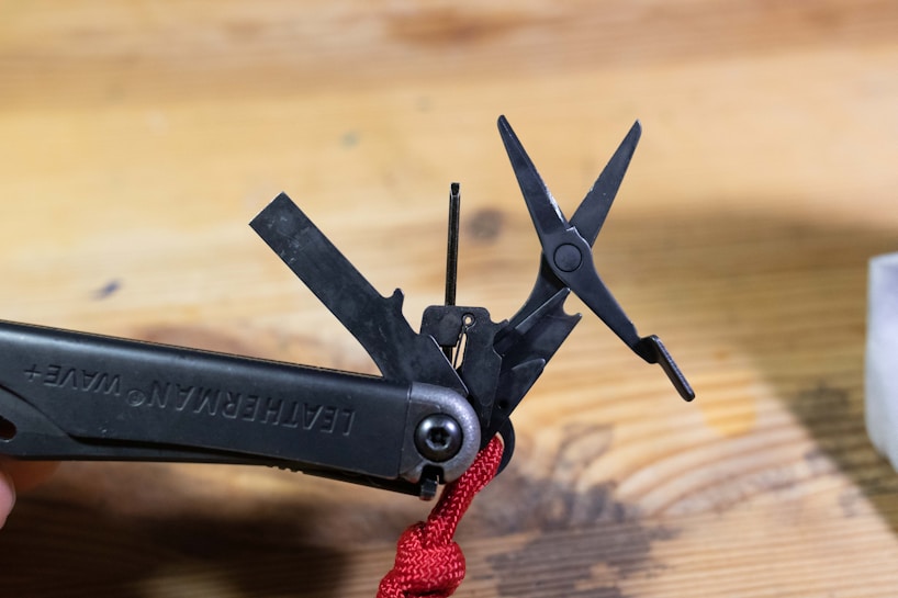 A multi-tool with a partially open pair of scissors and several other tools is shown on a wooden surface. One of the tools has a red cord attached to it. The name 'Leatherman Wave+' is visible on the handle.