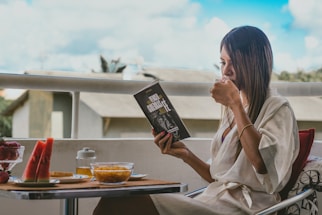 A person sits on a balcony, engrossed in reading a book titled 'Vencer Cuesta'. They are dressed in a white robe and holding a cup, possibly of tea or coffee. The small table in front is set with breakfast items including watermelon slices, a bowl of cereal, a jar of honey, and a dish with strawberries. The background shows an outdoor view with buildings and a bright, partly cloudy sky.