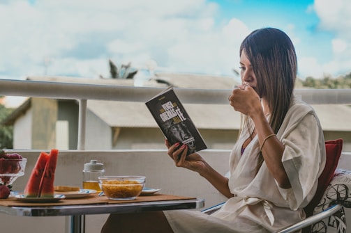 A person sits on a balcony, engrossed in reading a book titled 'Vencer Cuesta'. They are dressed in a white robe and holding a cup, possibly of tea or coffee. The small table in front is set with breakfast items including watermelon slices, a bowl of cereal, a jar of honey, and a dish with strawberries. The background shows an outdoor view with buildings and a bright, partly cloudy sky.