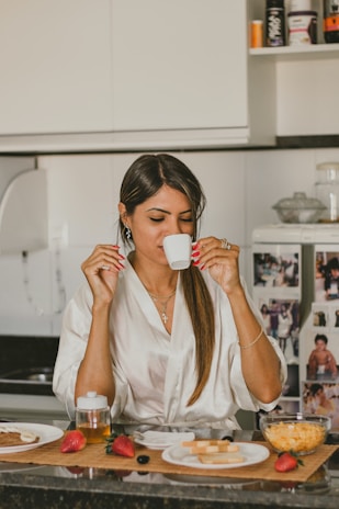 A woman in a kitchen enjoys a hot beverage from a small white cup. She is wearing a white robe and has long, straight dark hair. In front of her on a wooden mat are various breakfast items, including strawberries, a jar of honey, a plate with sliced cheese, and a bowl of cereal.
