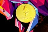 A gold coin featuring the Ethereum logo and name is set against a vibrant, geometric background with bold angular shapes in shades of purple, pink, orange, and blue.
