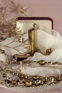 An ornate lighter rests in an open velvet-lined box, surrounded by delicate fabric. The lighter is accompanied by gold and silver-toned decorative beads and dry twigs, creating a serene and vintage aesthetic.