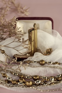 An ornate lighter rests in an open velvet-lined box, surrounded by delicate fabric. The lighter is accompanied by gold and silver-toned decorative beads and dry twigs, creating a serene and vintage aesthetic.