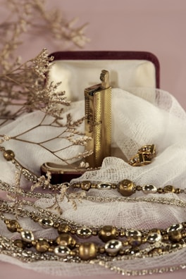 An ornate lighter rests in an open velvet-lined box, surrounded by delicate fabric. The lighter is accompanied by gold and silver-toned decorative beads and dry twigs, creating a serene and vintage aesthetic.