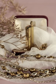 An ornate lighter rests in an open velvet-lined box, surrounded by delicate fabric. The lighter is accompanied by gold and silver-toned decorative beads and dry twigs, creating a serene and vintage aesthetic.