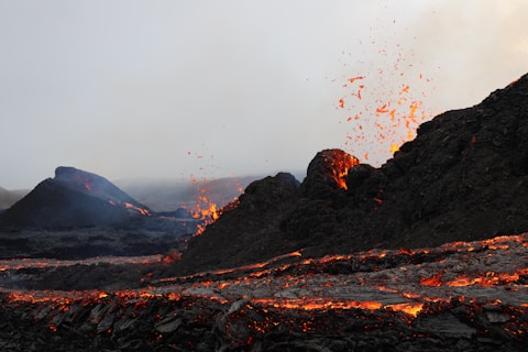Molten lava spews from a volcanic eruption, with fiery red and orange lava contrasting against dark volcanic rocks. The sky is overcast, adding a dramatic and intense atmosphere.