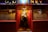 A man is entering a warmly lit, traditional pub through a wooden door. The exterior features red paneling and gold lettering spelling 'G. Delaney' at the top. Frosted glass windows display the words 'Delaney' and 'Bar & Lounge'.