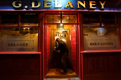 A man is entering a warmly lit, traditional pub through a wooden door. The exterior features red paneling and gold lettering spelling 'G. Delaney' at the top. Frosted glass windows display the words 'Delaney' and 'Bar & Lounge'.