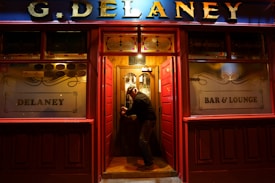 A man is entering a warmly lit, traditional pub through a wooden door. The exterior features red paneling and gold lettering spelling 'G. Delaney' at the top. Frosted glass windows display the words 'Delaney' and 'Bar & Lounge'.