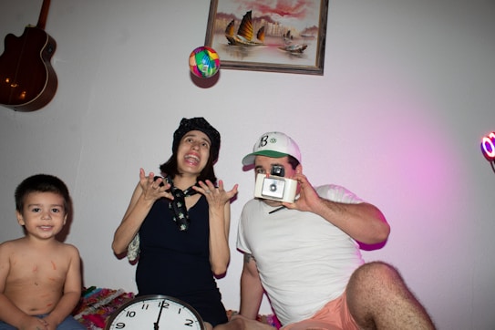 A family scene with three people is depicted, including a child, a woman, and a man sitting on a bed or couch. The woman appears to be tossing a colorful ball into the air while enthusiastically gesturing with her hands. The man, wearing a white shirt and a cap, is holding an old-fashioned camera and posing as if taking a picture. The child is smiling and sitting calmly with some playful markings on his body. On the wall behind them, there's a framed picture of sailboats and a guitar hung up.