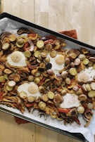 A baking sheet filled with roasted vegetables and eggs sits atop a wooden table. The vegetables include sliced potatoes, bell peppers, and onions, all seasoned with herbs. Several fried eggs are nestled among the vegetables, their yolks still intact.