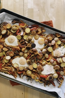 A baking sheet filled with roasted vegetables and eggs sits atop a wooden table. The vegetables include sliced potatoes, bell peppers, and onions, all seasoned with herbs. Several fried eggs are nestled among the vegetables, their yolks still intact.