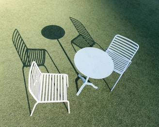 Two white metal chairs with horizontal slats are positioned outdoors on a green, textured surface. A round white metal table is placed between the chairs. The scene is brightly lit, casting distinct shadows of the chairs and table.