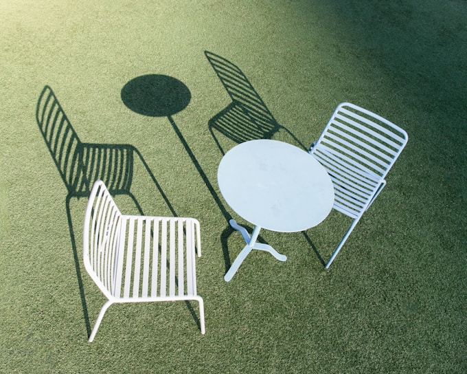 Two white metal chairs with horizontal slats are positioned outdoors on a green, textured surface. A round white metal table is placed between the chairs. The scene is brightly lit, casting distinct shadows of the chairs and table.