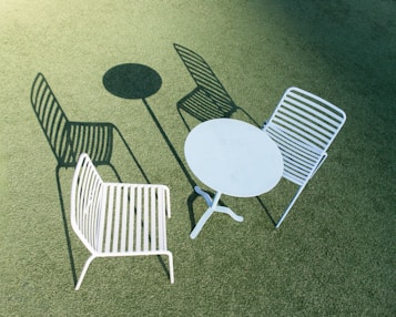 Two white metal chairs with horizontal slats are positioned outdoors on a green, textured surface. A round white metal table is placed between the chairs. The scene is brightly lit, casting distinct shadows of the chairs and table.