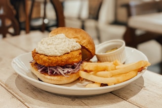 A plate containing a sandwich with a crispy fried patty topped with a creamy sauce, placed on a bed of shredded cabbage in a bun. Beside the sandwich, there are several golden French fries. A small white ramekin with a pickle or similar garnish is also present on the plate. The setting appears to be a cozy indoor dining area with wooden furniture.
