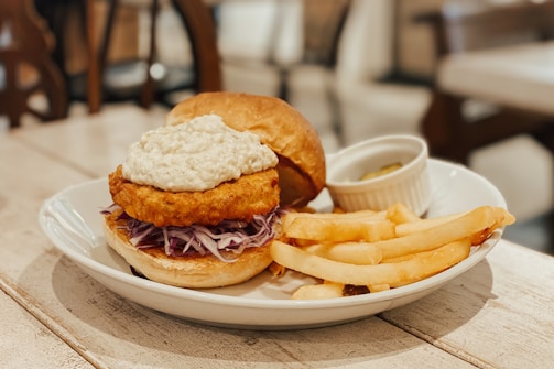 A plate containing a sandwich with a crispy fried patty topped with a creamy sauce, placed on a bed of shredded cabbage in a bun. Beside the sandwich, there are several golden French fries. A small white ramekin with a pickle or similar garnish is also present on the plate. The setting appears to be a cozy indoor dining area with wooden furniture.