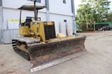 A yellow bulldozer is parked on a construction site. The machine has a large, rusty metal blade in the front and caterpillar tracks. It is positioned on a concrete surface near a building with large windows. Surrounding the site are various construction materials and equipment, with some greenery and vehicles visible in the background.