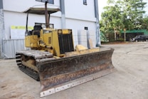 A yellow bulldozer is parked on a construction site. The machine has a large, rusty metal blade in the front and caterpillar tracks. It is positioned on a concrete surface near a building with large windows. Surrounding the site are various construction materials and equipment, with some greenery and vehicles visible in the background.