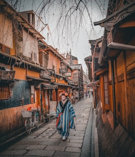 A person wearing traditional Japanese attire walks down a narrow, cobblestone street lined with traditional wooden buildings. The street is empty except for a bicycle parked on the side. Overhead, bare tree branches hang down against a cloudy sky, adding to the serene and historic atmosphere.