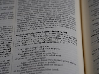 A close-up of a Bible page highlighting a passage from the book of Isaiah, specifically Isaiah prophesying Sennacherib's fall. The text is clearly focused and well lit, with adjacent text pages slightly blurred due to depth of field.