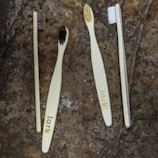 Four toothbrushes with bamboo handles and varying bristle colors are arranged on a textured stone surface. The handles are engraved with the word 'lark', and the bristle colors include brown, black, beige, and white.