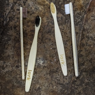 Four toothbrushes with bamboo handles and varying bristle colors are arranged on a textured stone surface. The handles are engraved with the word 'lark', and the bristle colors include brown, black, beige, and white.