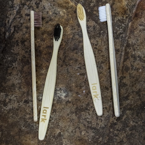 Four toothbrushes with bamboo handles and varying bristle colors are arranged on a textured stone surface. The handles are engraved with the word 'lark', and the bristle colors include brown, black, beige, and white.